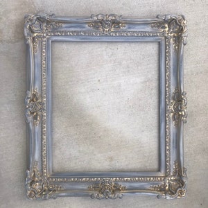 Puede incluir: Un marco de imagen adornado en gris y dorado con un dise&ntilde;o decorativo. El marco est&aacute; hecho de madera y tiene un acabado desgastado.