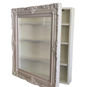 May include: A wall-mounted display cabinet featuring a light beige, ornate, carved frame with a glass front door. The interior contains three horizontal white shelves. The side of the cabinet is visible, showing a plain white rectangular structure.