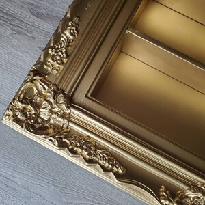 May include: A gold-coloured picture frame with ornate detailing and a decorative, raised design. The frame has two shelves inside.