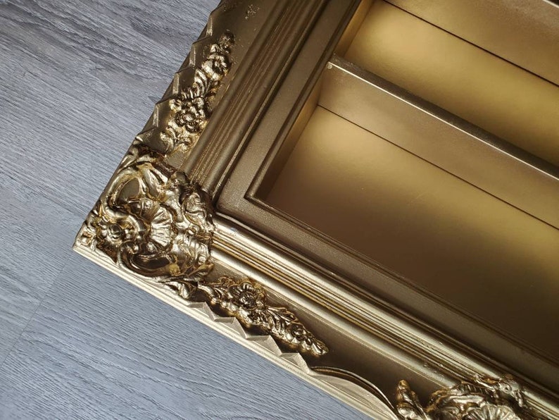 May include: A gold-coloured picture frame with ornate detailing and a decorative, raised design. The frame has two shelves inside.