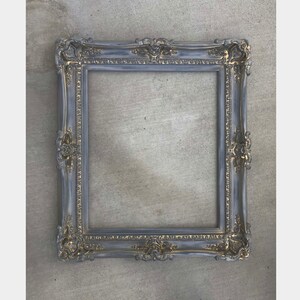 Puede incluir: Un marco de imagen ornamentado en gris y dorado con un dise&ntilde;o decorativo y detallado. El marco est&aacute; hecho de madera y tiene un acabado desgastado.