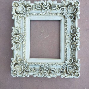 May include: A white ornate picture frame with a distressed finish. The frame has a detailed design with intricate carvings and a slightly aged look.
