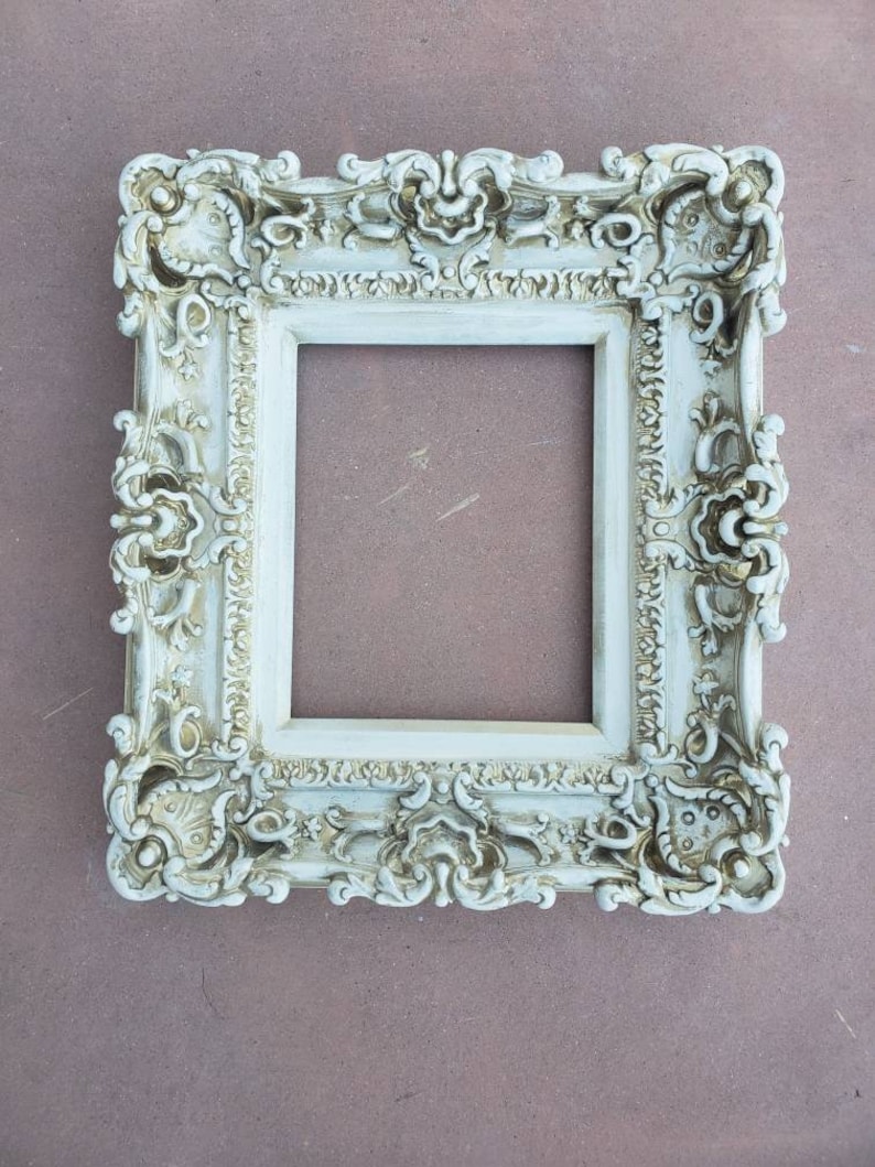May include: A white ornate picture frame with a distressed finish. The frame has a detailed design with intricate carvings and a slightly aged look.