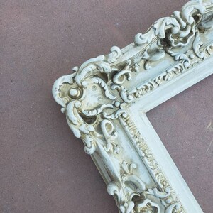 May include: A white ornate picture frame with gold accents. The frame has a detailed, intricate design with a floral motif.