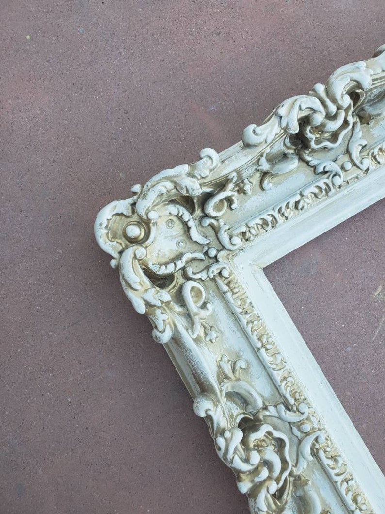 May include: A white ornate picture frame with gold accents. The frame has a detailed, intricate design with a floral motif.