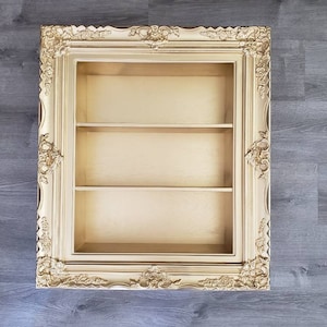 May include: Gold ornate picture frame with three shelves inside. The frame is a decorative display shelf.
