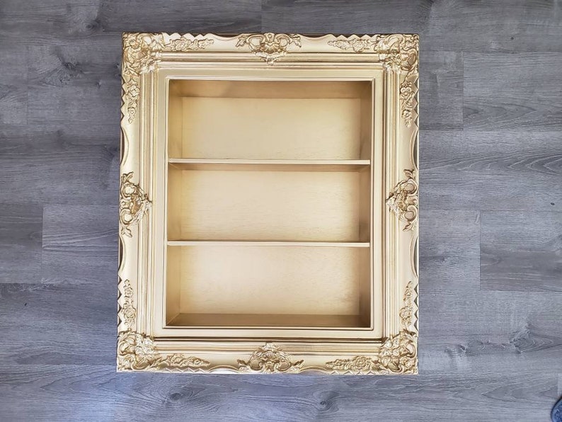 May include: Gold ornate picture frame with three shelves inside. The frame is a decorative display shelf.