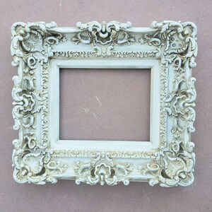 May include: A white ornate picture frame with a highly detailed design. The frame has a rectangular shape and a raised edge around the opening.