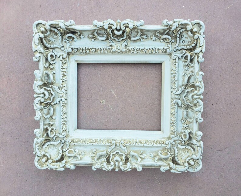 May include: A white ornate picture frame with a highly detailed design. The frame has a rectangular shape and a raised edge around the opening.