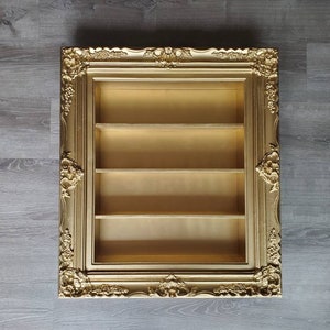 May include: A gold-coloured picture frame with ornate detailing, featuring three shelves inside. The frame is empty and ready to be decorated.