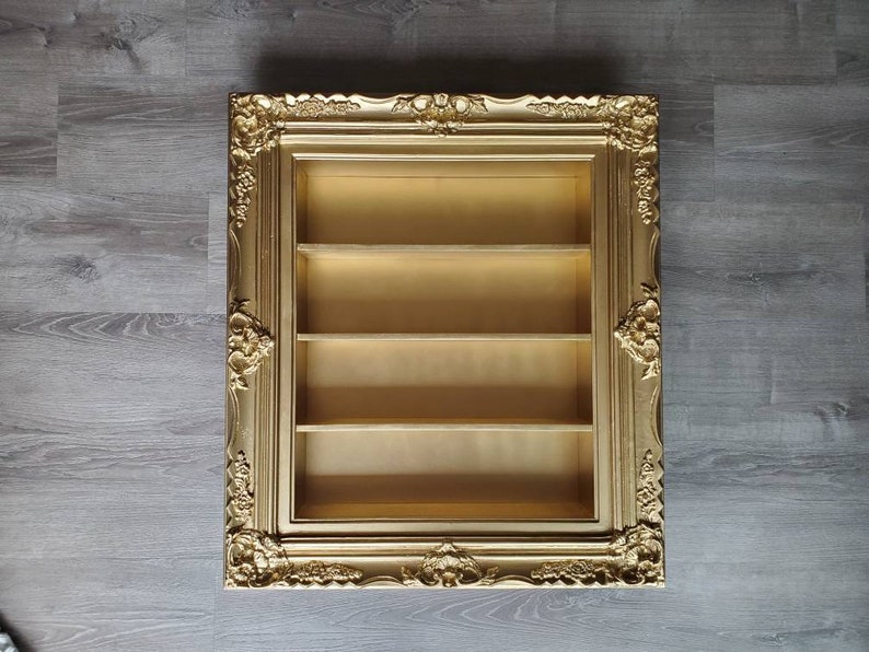 May include: A gold-coloured picture frame with ornate detailing, featuring three shelves inside. The frame is empty and ready to be decorated.