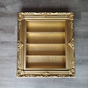 May include: A gold-coloured picture frame with ornate detailing and three shelves inside. The frame is empty.
