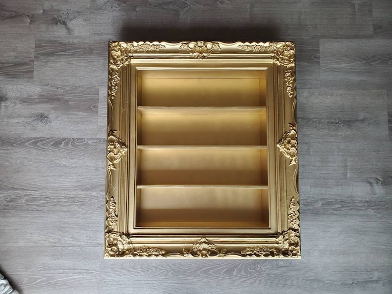May include: A gold-coloured picture frame with ornate detailing and three shelves inside. The frame is empty.