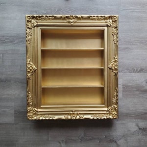 May include: A gold-coloured picture frame with ornate detailing, featuring a three-shelf display case inside.