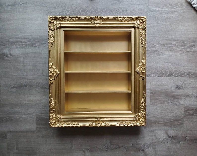 May include: A gold-coloured picture frame with ornate detailing, featuring a three-shelf display case inside.