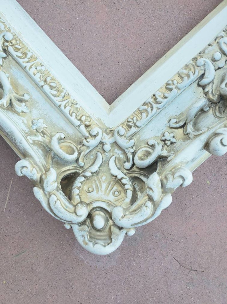 May include: A white ornate picture frame with a gold-toned design. The frame has a decorative, ornate design with a raised, textured surface.