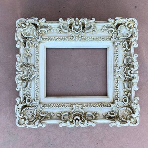 May include: A white ornate picture frame with a highly detailed design. The frame has a raised edge and a smooth, flat surface.