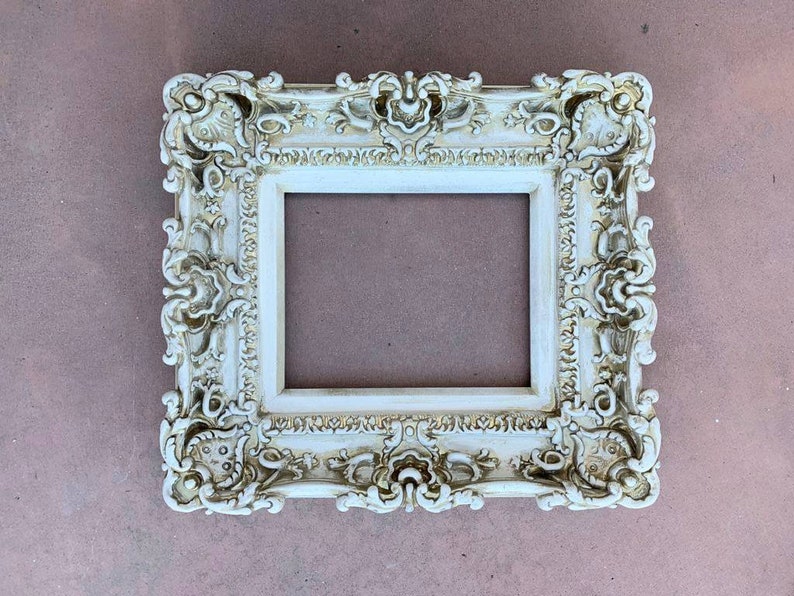 May include: A white ornate picture frame with a highly detailed design. The frame has a raised edge and a smooth, flat surface.