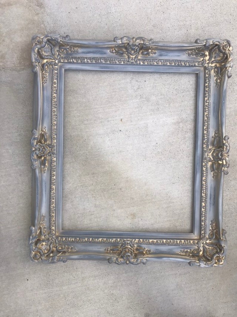 Puede incluir: Un marco de imagen adornado en gris y dorado con un acabado desgastado. El marco tiene un dise&ntilde;o decorativo con detalles intrincados.