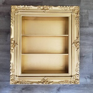May include: A gold-colored picture frame with a shelf inside. The frame has ornate details and is made of wood.
