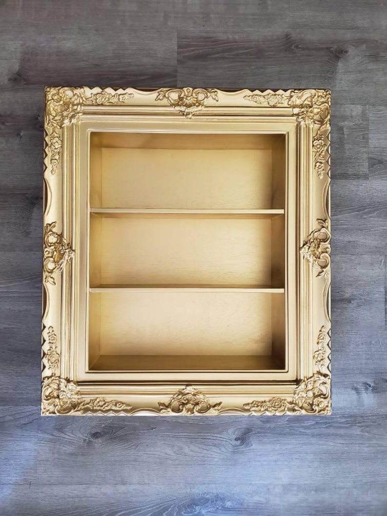 May include: A gold-colored picture frame with a shelf inside. The frame has ornate details and is made of wood.