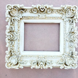 May include: A white ornate picture frame with a highly detailed, intricate design. The frame is made of a smooth, white material and has a slightly aged appearance.