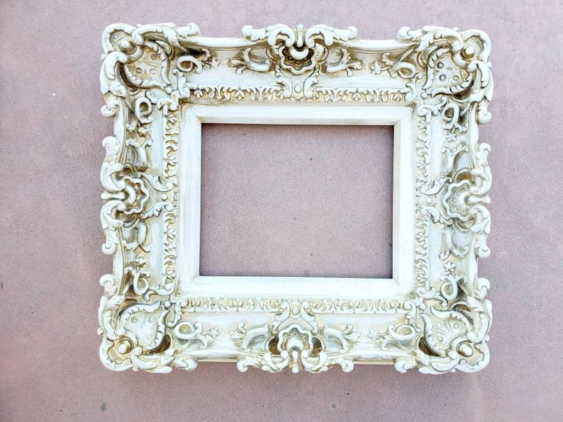 May include: A white ornate picture frame with a highly detailed, intricate design. The frame is made of a smooth, white material and has a slightly aged appearance.