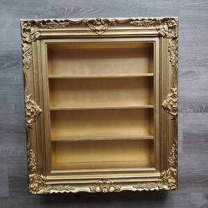 May include: A gold-coloured picture frame with a decorative ornate design. The frame has three shelves inside.