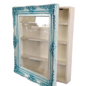 May include: A wall-mounted display cabinet featuring a white interior with three horizontal shelves. The front is a hinged door framed in ornate, textured moulding painted in a distressed white and bright turquoise blue finish with glass.