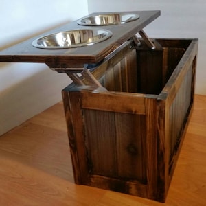 May include: A wooden pet food and water bowl stand with a hinged lid that lifts to reveal a storage compartment. The stand is stained a dark brown colour and has a rustic appearance.