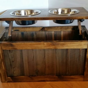 May include: A wooden dog food and water bowl stand with a storage compartment. The stand has two stainless steel bowls and is made of dark brown wood.