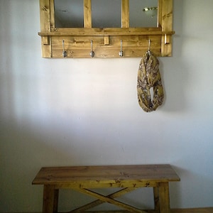 May include: A wooden entryway bench with a shelf and mirror above. The shelf has five hooks for hanging coats or bags. The bench is made of natural wood with a light brown stain.