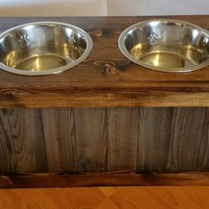 May include: A wooden dog bowl stand with two stainless steel bowls. The stand has a rustic brown finish and a grey wood panel on the front.