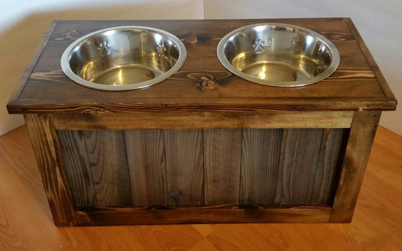 May include: A wooden dog bowl stand with two stainless steel bowls. The stand has a rustic brown finish and a grey wood panel on the front.
