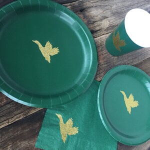 May include: Three green paper plates with gold glitter duck silhouettes. The plates are on a wooden surface with a green napkin.