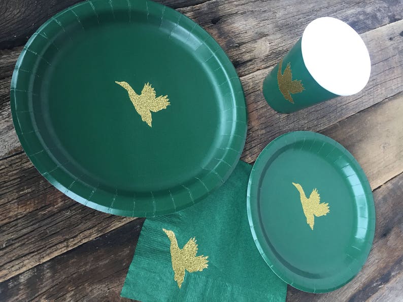 May include: Three green paper plates with gold glitter duck silhouettes. The plates are on a wooden surface with a green napkin.