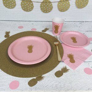 May include: A pink and gold pineapple themed party table setting with a gold glitter charger, a pink paper plate, a pink paper napkin, a pink paper cup, and a gold fork. The table is decorated with gold pineapple cutouts.
