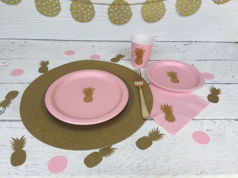 May include: A pink and gold pineapple themed party table setting with a gold glitter charger, a pink paper plate, a pink paper napkin, a pink paper cup, and a gold fork. The table is decorated with gold pineapple cutouts.