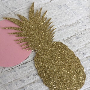 May include: Gold glitter pineapple cutout with a pink circle behind it. The pineapple is on a white wooden surface.