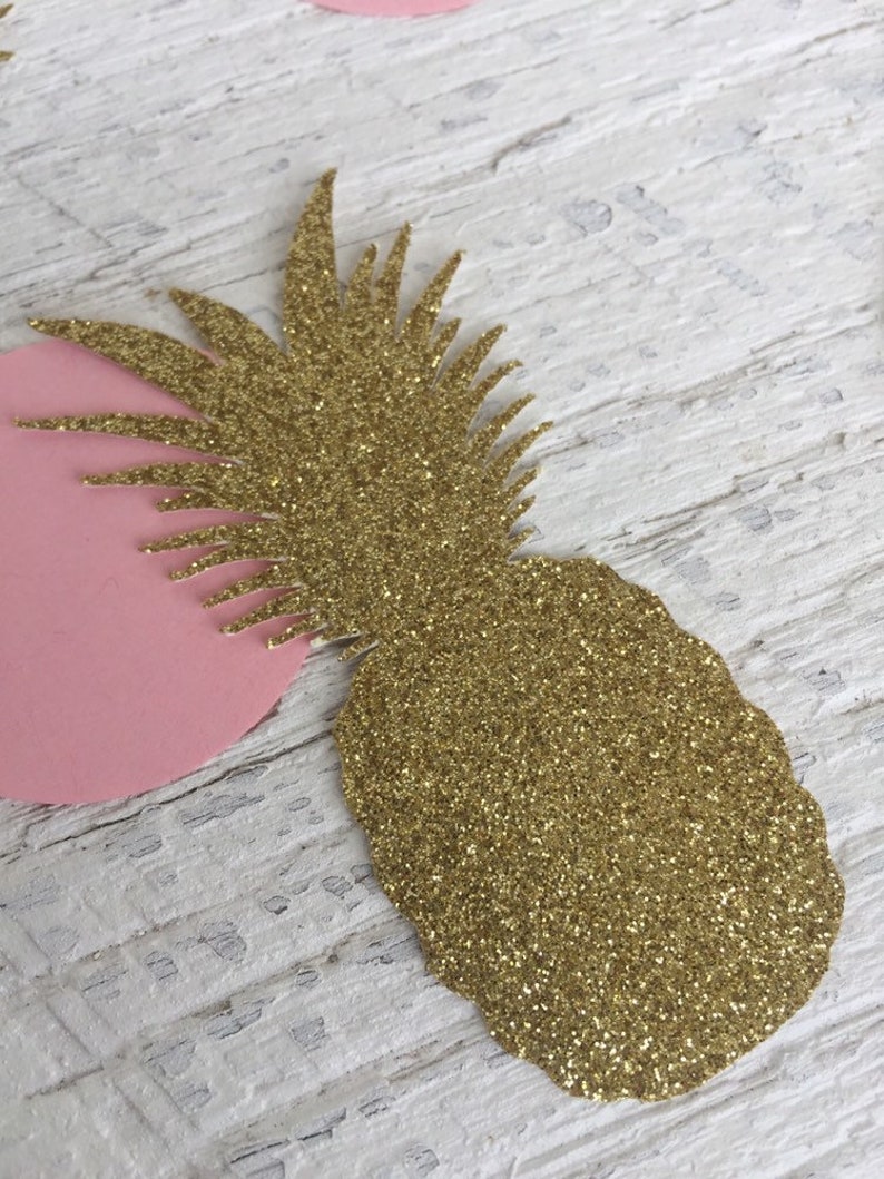 May include: Gold glitter pineapple cutout with a pink circle behind it. The pineapple is on a white wooden surface.