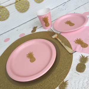 May include: Pink and gold pineapple themed party supplies. A pink paper plate with a gold pineapple design sits on a gold glitter charger. A pink paper cup with a gold pineapple design is next to the plate. A gold fork is on the table.