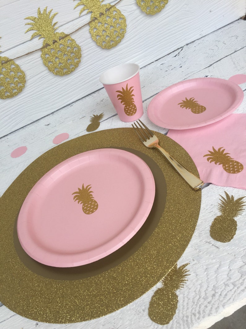May include: Pink and gold pineapple themed party supplies. A pink paper plate with a gold pineapple design sits on a gold glitter charger. A pink paper cup with a gold pineapple design is next to the plate. A gold fork is on the table.