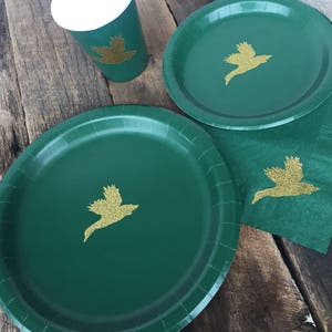 May include: Green paper plates and cups with gold glitter bird designs. The plates are round and have a scalloped edge. The cups are white with a green rim and bottom.