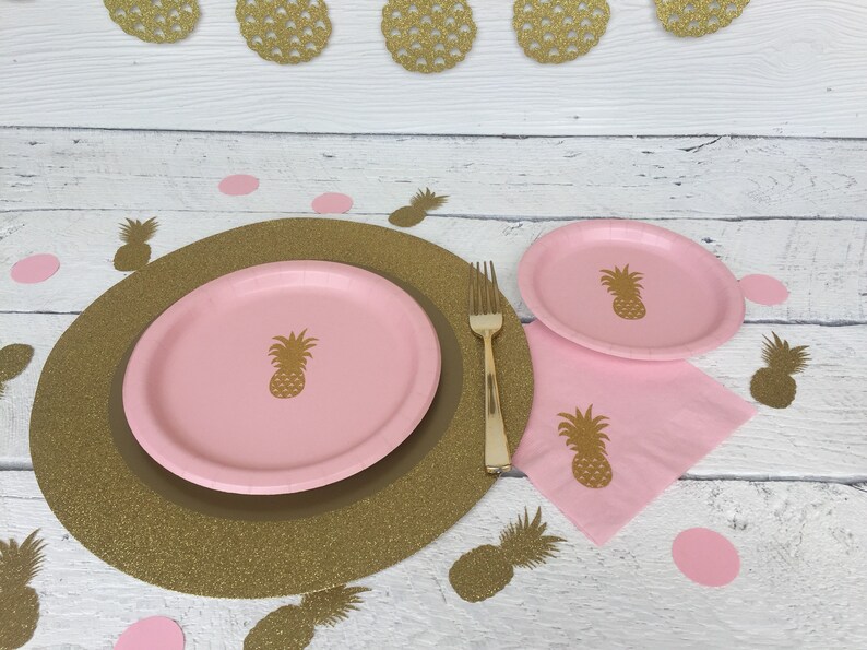 May include: A pink and gold pineapple themed table setting with a gold glitter charger, a pink dinner plate, a pink dessert plate, a pink napkin, and a gold fork. The table setting is decorated with pink and gold pineapple cutouts.