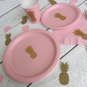 May include: Pink paper plates and cups with gold glitter pineapple designs. The plates and cups are on a white wooden surface with pink and gold glitter confetti.