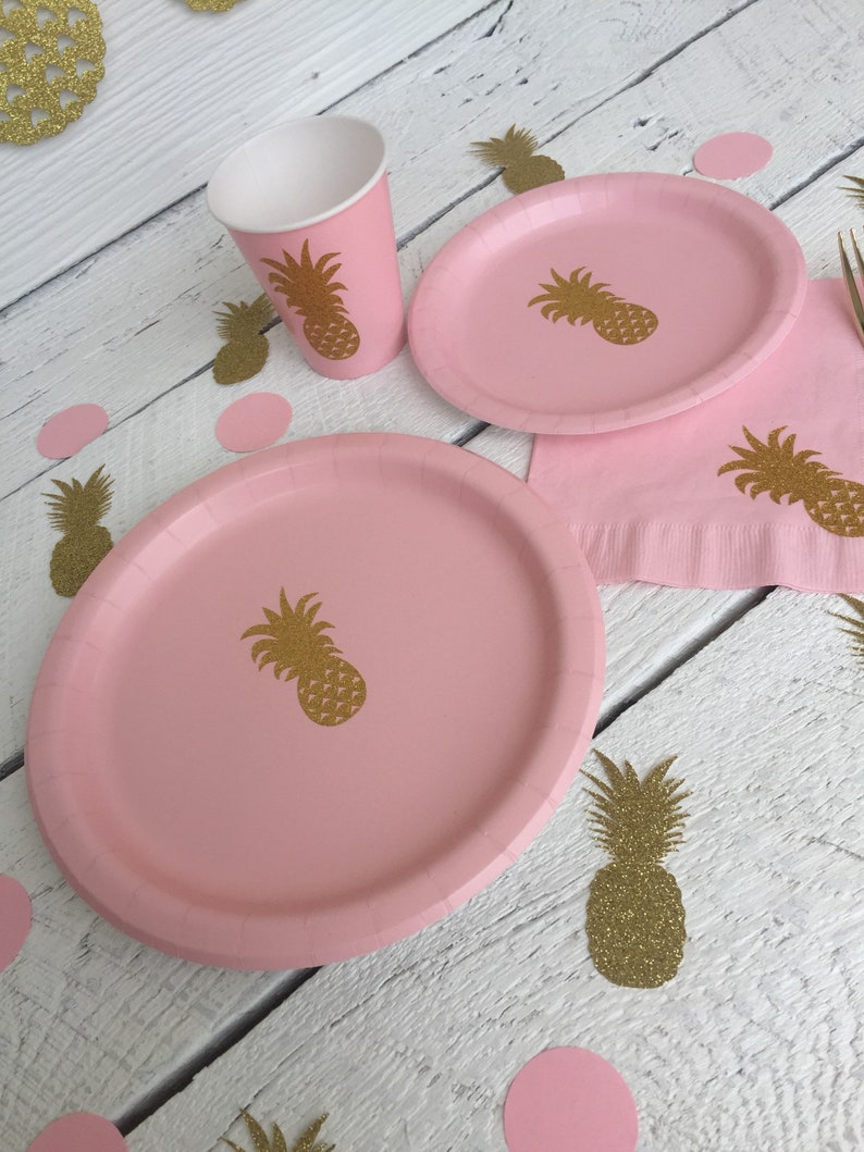 May include: Pink paper plates and cups with gold glitter pineapple designs. The plates and cups are on a white wooden surface with pink and gold glitter confetti.