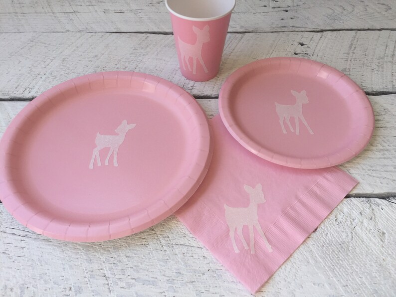 May include: Pink paper plates and cups with a white silhouette of a deer. Pink paper napkins with a white silhouette of a deer.