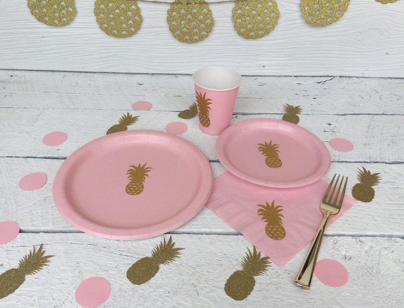 May include: Pink paper plates, cups, and napkins with a pineapple design. Gold glitter pineapple confetti is scattered on a white wooden surface.