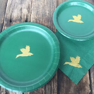 May include: Three green paper plates with gold flying duck silhouettes. The plates are on a wooden surface.