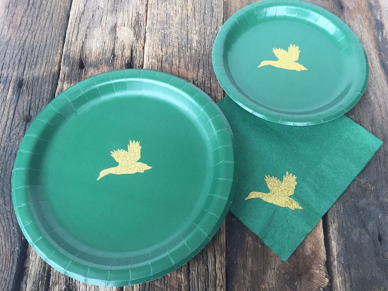 May include: Three green paper plates with gold flying duck silhouettes. The plates are on a wooden surface.